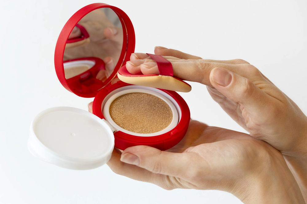 A person holds a red compact case with a mirror and cushion foundation, using a make-up puff applicator to pick up the product with their fingers.