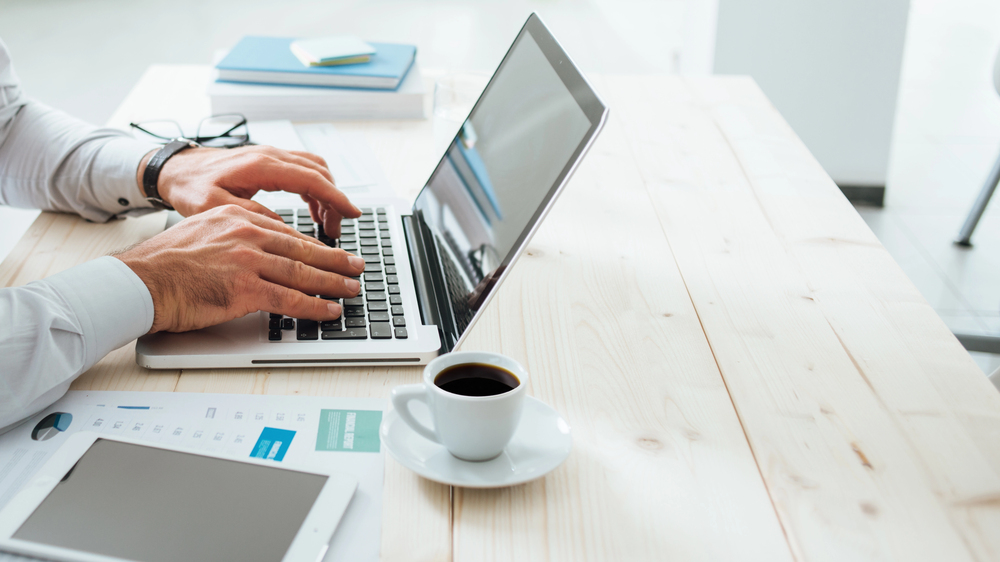 A person types on a laptop at a wooden desk with documents, a tablet, stacked books, and a cup of coffee nearby in a bright, modern office setting.