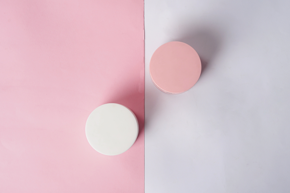 Two round containers, one white and one pink, placed side by side on a background split vertically into pink (left) and white (right), each container matching the colour of the background on the opposite side.