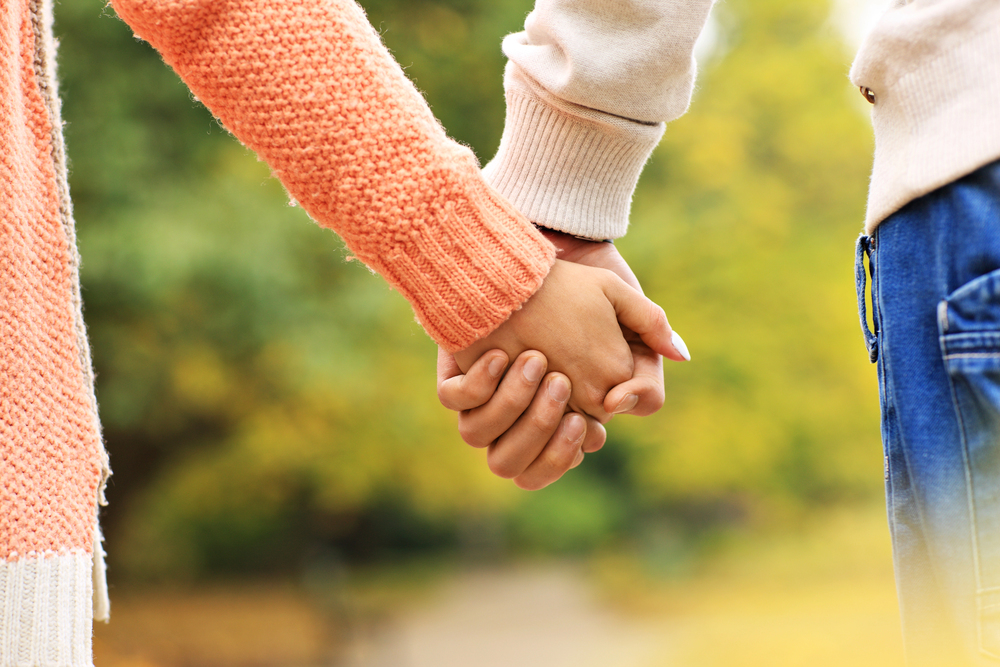 Two people holding hands outdoors, one wearing an orange knitted jumper and the other wearing a light-coloured jumper and blue jeans, with a blurred background of green and yellow foliage.