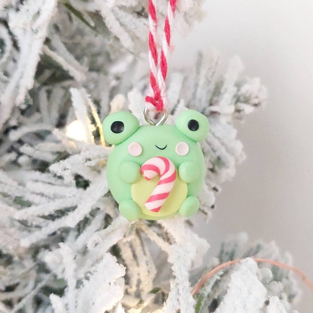 A cute green frog ornament holding a red and white candy cane hangs on a snow-covered Christmas tree branch with a string.