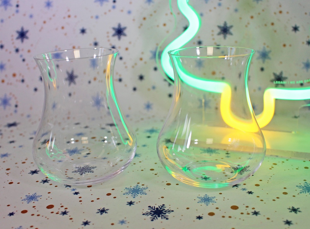 Two clear, curved glasses sit on a surface decorated with blue snowflakes. In the background, there is a wavy neon light glowing green and yellow, creating a festive and colourful ambience.