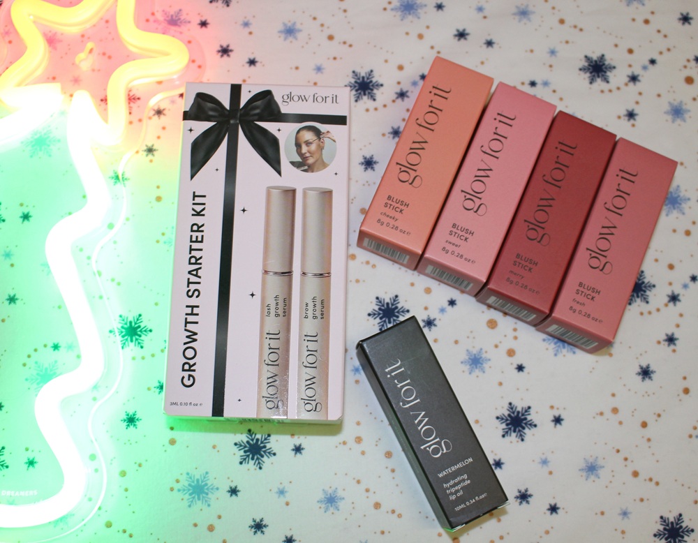 Glow For It beauty products on a snowflake-patterned surface with a neon light. Items include a Growth Starter Kit, four boxed blushers in pink shades, and one black boxed product.
