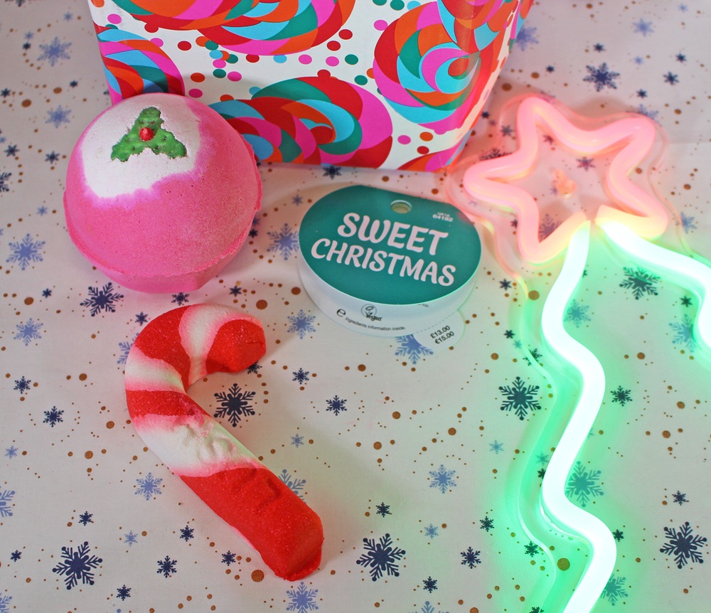 A festive scene with a pink bath bomb, a Sweet Christmas tin, a red and white candy cane, and a glowing neon star wand on snowflake-patterned paper, next to a colourful gift box.