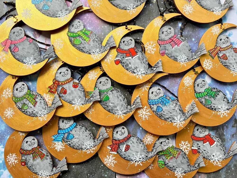 Several illustrated ornaments of smiling seals in colourful scarves and hats rest on golden crescent moons, surrounded by white snowflakes against a starry background. Each crescent features a different festive colour.