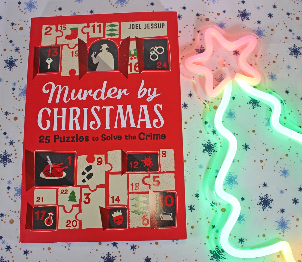 A book titled Murder by Christmas: 25 Puzzles to Solve the Crime by Joel Jessup rests on festive, snowflake-patterned paper next to a neon light shaped like a Christmas tree with a pink star.