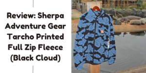 A blue and black cloud-patterned fleece jacket hangs on a wooden hanger outdoors by a pond. Text on the left reads: “Review: Sherpa Adventure Gear Tarcho Printed Full Zip Fleece (Black Cloud).”.
