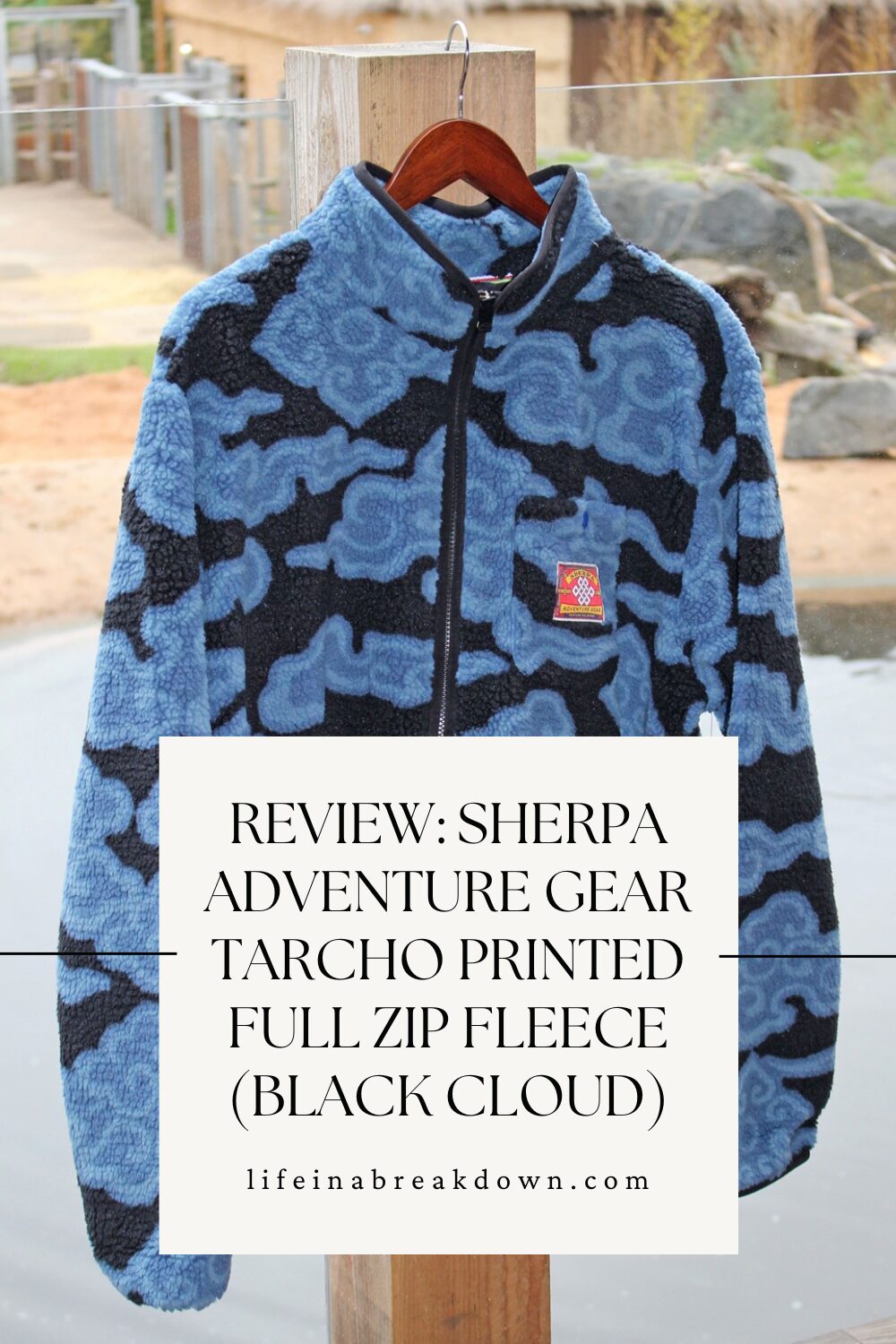 A blue and black patterned fleece jacket hangs on a wooden hanger outdoors, with a review text overlay about the Sherpa Adventure Gear Tarcho Printed Full Zip Fleece (Black Cloud).