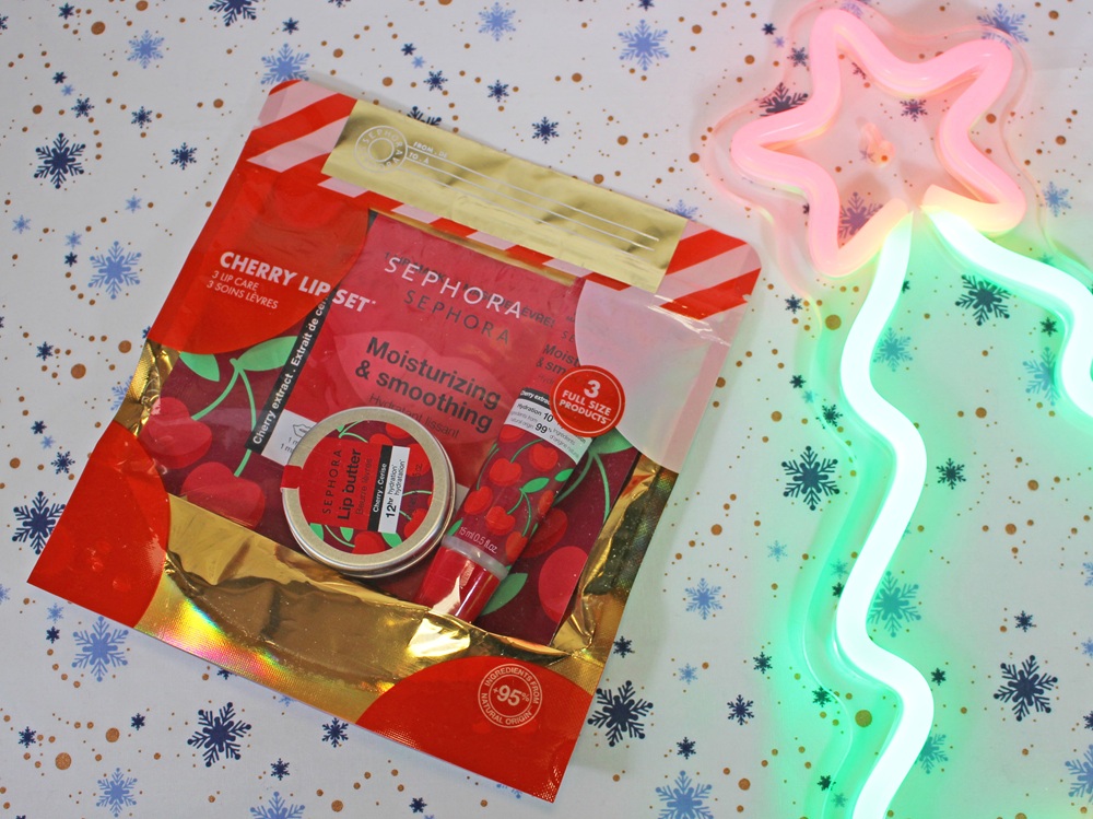 A Sephora Cherry Lip Set with balm and scrub in red packaging sits on a snowflake-patterned surface next to a pink and green neon light shaped like a star and tree.