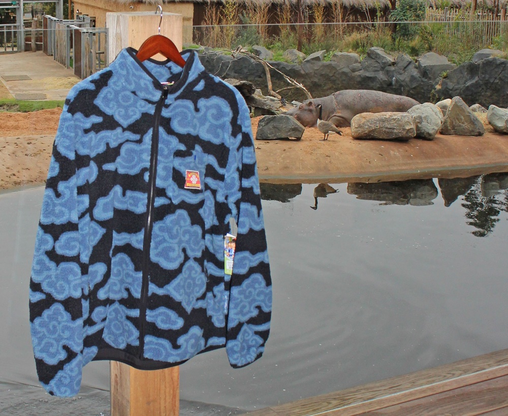 A blue and black patterned fleece jacket hangs on a wooden post in front of a hippo enclosure at a zoo, with a hippo and birds visible near the water in the background.
