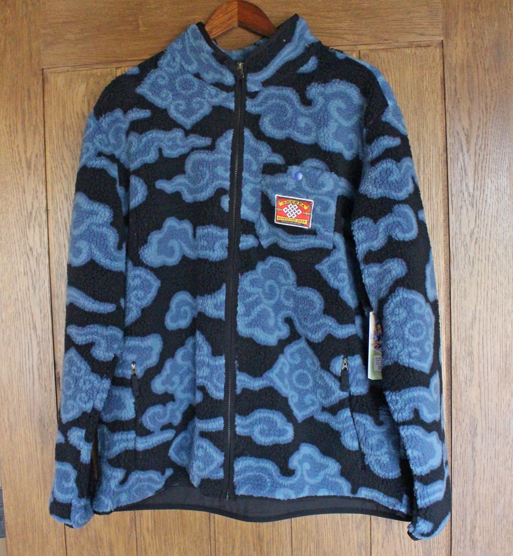 A blue and black fleece jacket with a cloud pattern hangs on a wooden hanger against a wooden door. The jacket has a front zip, two zipped pockets, and a red patterned patch on the chest.