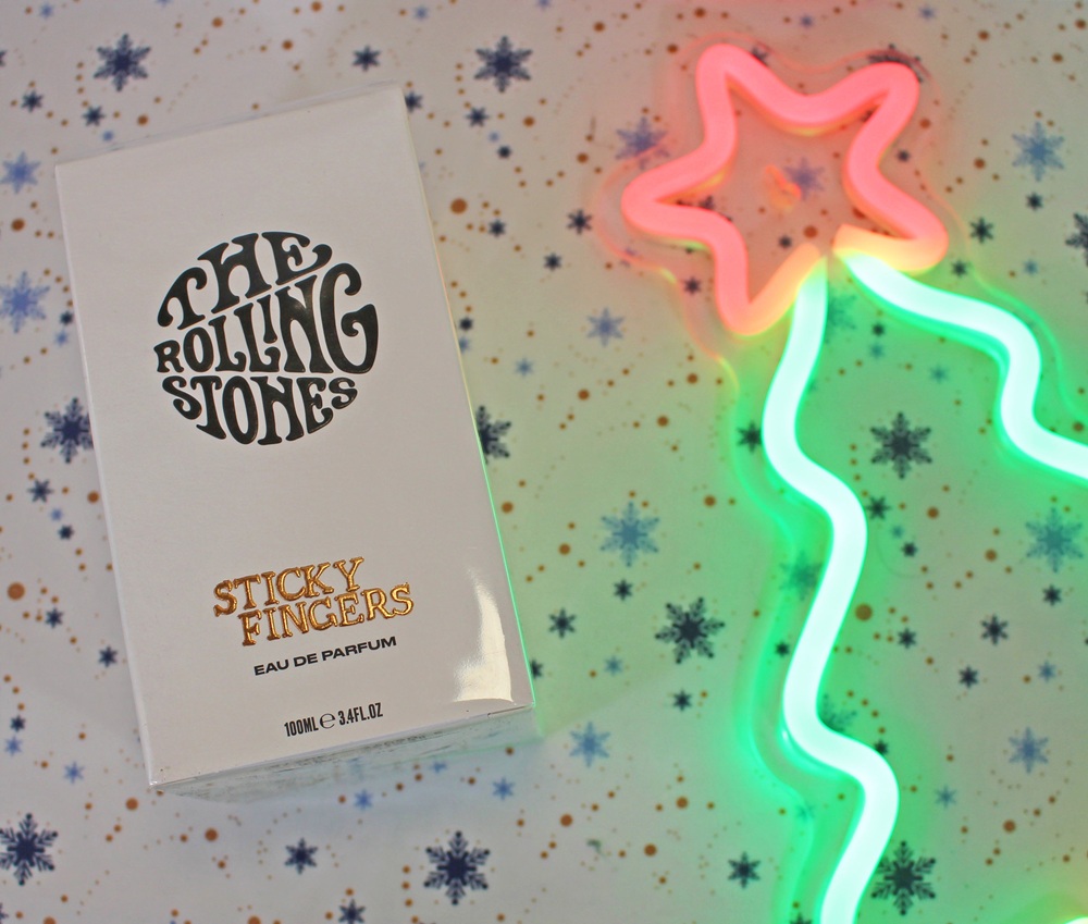 A box of The Rolling Stones Sticky Fingers Eau de Parfum sits on a festive snowflake-patterned background beside a neon pink and green star-shaped light.