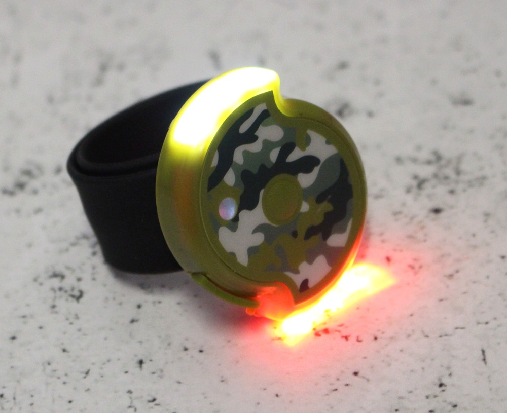 A small round wearable device, the Twistii Torch, features a camouflage pattern and glows with yellow and red LED lights, attached to a black strap and placed on a speckled white surface.