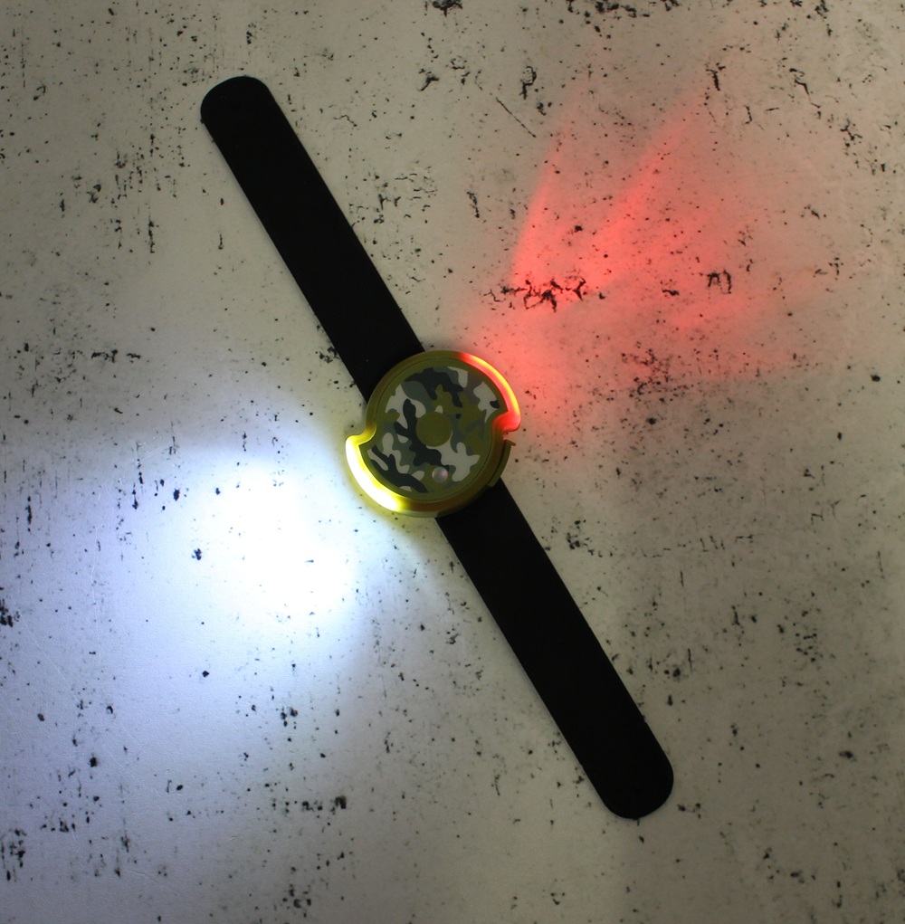 A Twistii Torch wristband with a camouflage-patterned circular centre emits red and white lights onto a speckled, greyish surface.