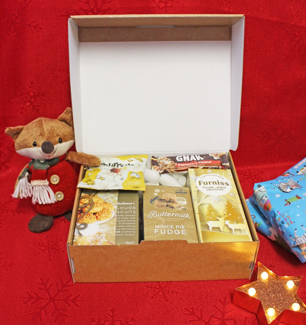 A festive gift box filled with various snacks and treats sits open on a red background. A soft toy fox is beside the box, with wrapped presents and a lit star decoration nearby.