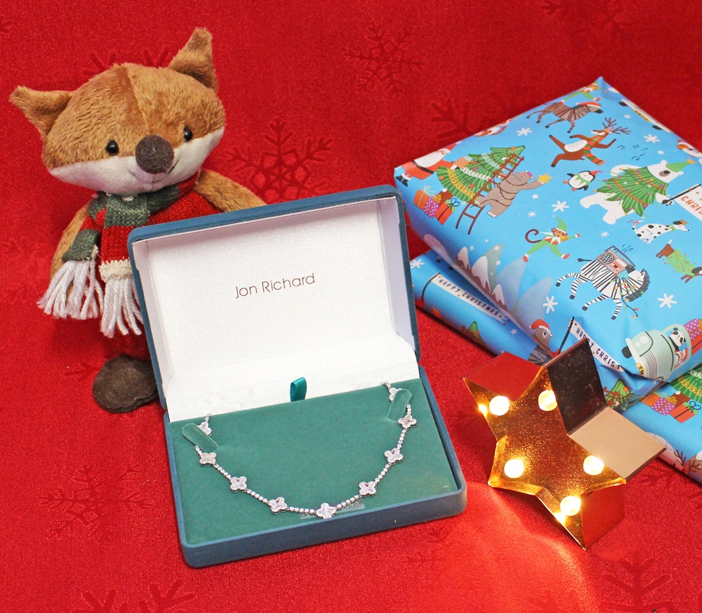 A plush fox, a Jon Richard necklace in a gift box, a star-shaped light, and two wrapped presents with festive designs on a red holiday background.