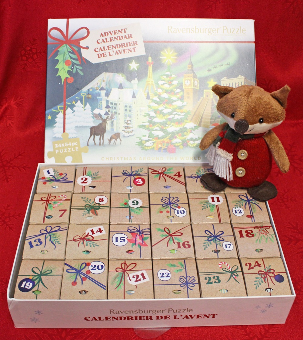 A Ravensburger Puzzle advent calendar with 24 numbered boxes, decorated with coloured bows and festive symbols, sits open. A plush fox wearing a red coat and scarf is perched on the edge of the box.