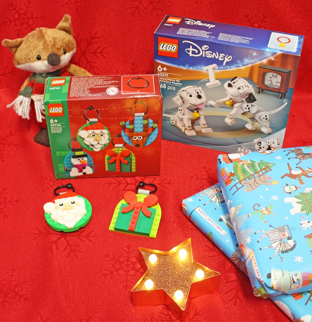 A festive scene with a plush fox, two LEGO sets (one Disney 101 Dalmatians, one Christmas), two LEGO holiday baubles, wrapped presents with festive paper, and a glowing star-shaped light on a red background.
