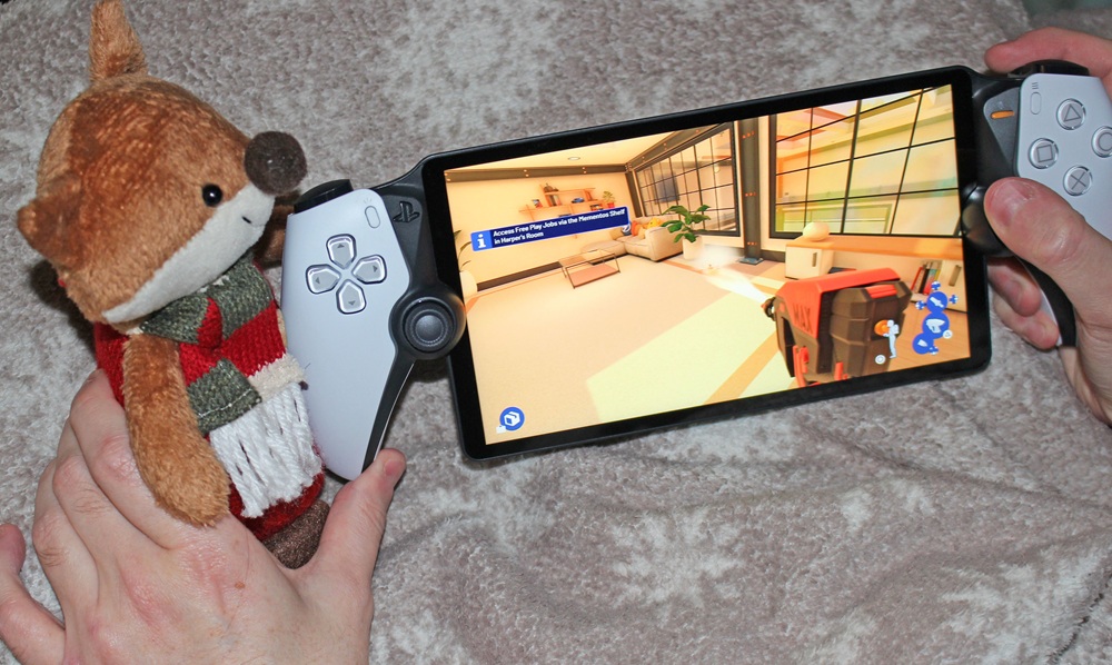 A person holds a tablet with game controllers attached, displaying a bright indoor game scene. A small plush fox toy wearing a scarf is held in one hand alongside the controller.