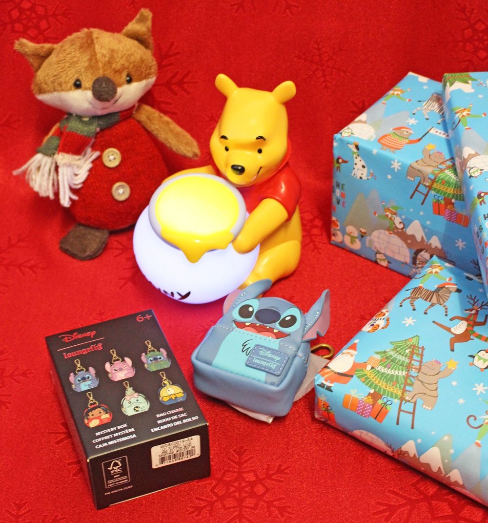A plush fox, a Winnie the Pooh toy holding a glowing pot, a box of Disney keyrings, a Stitch coin purse, and two wrapped presents are displayed on a red festive background.