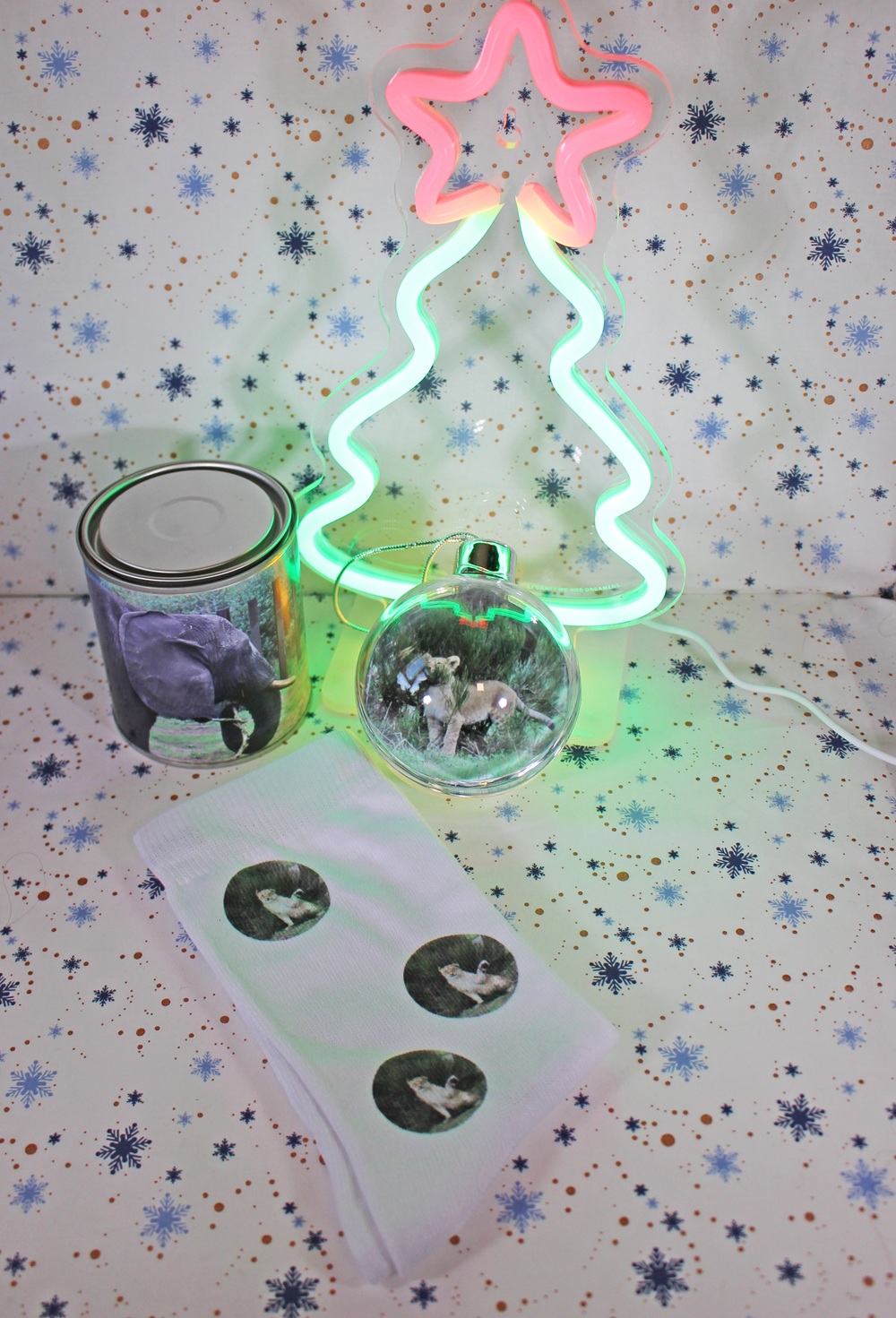 A glowing neon Christmas tree light, a tin with an animal photo label, a clear bauble with an animal image inside, and a white cloth with three animal stickers, all on a snowflake-patterned background.