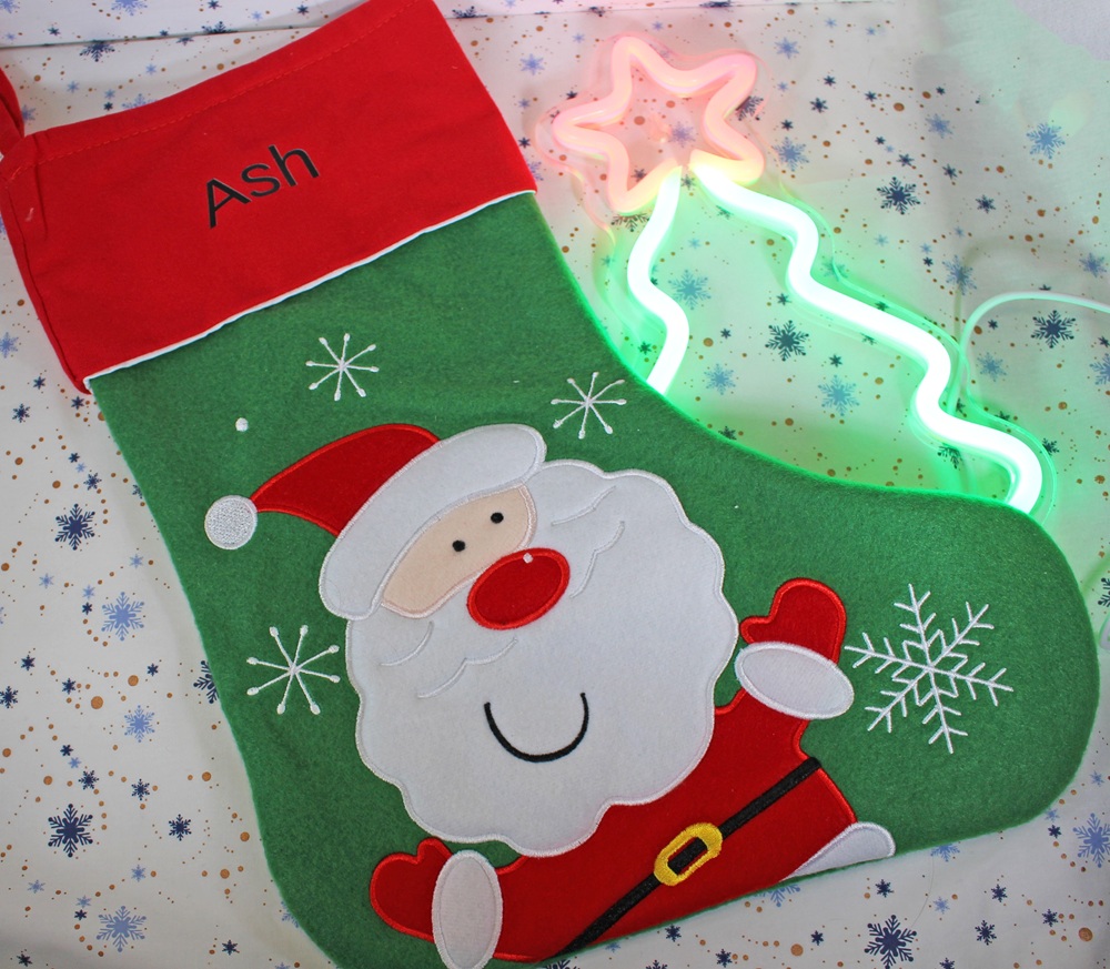 A green Christmas stocking with a red top labelled Ash features an appliqué of Father Christmas and white snowflakes. A neon light shaped like a Christmas tree glows beside it, on snowflake-patterned wrapping paper.