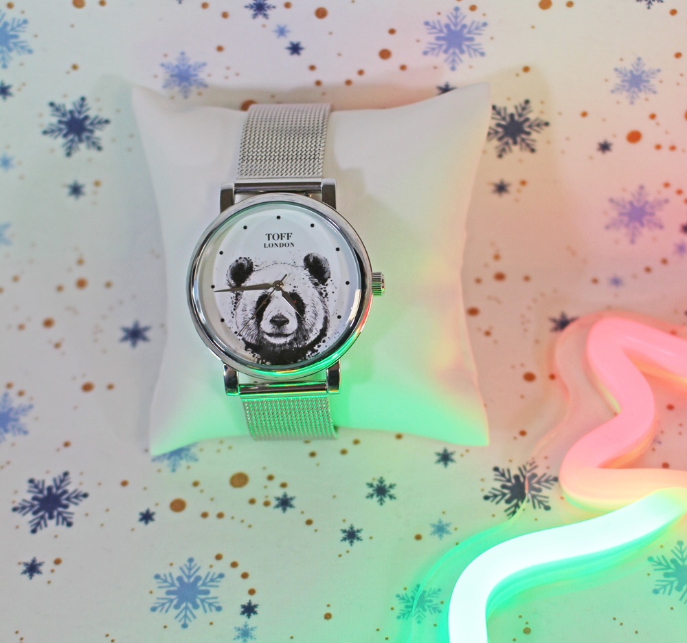A silver wristwatch with a panda face design is displayed on a white cushion. The background features snowflake patterns and colourful lights, creating a festive atmosphere.
