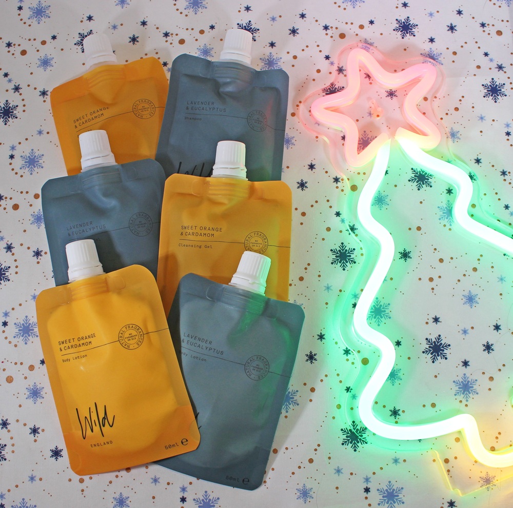 Five pouches of skincare products are arranged on a snowflake-patterned surface next to a glowing neon outline of a Christmas tree with a pink star on top. The pouches are in yellow and grey colours.