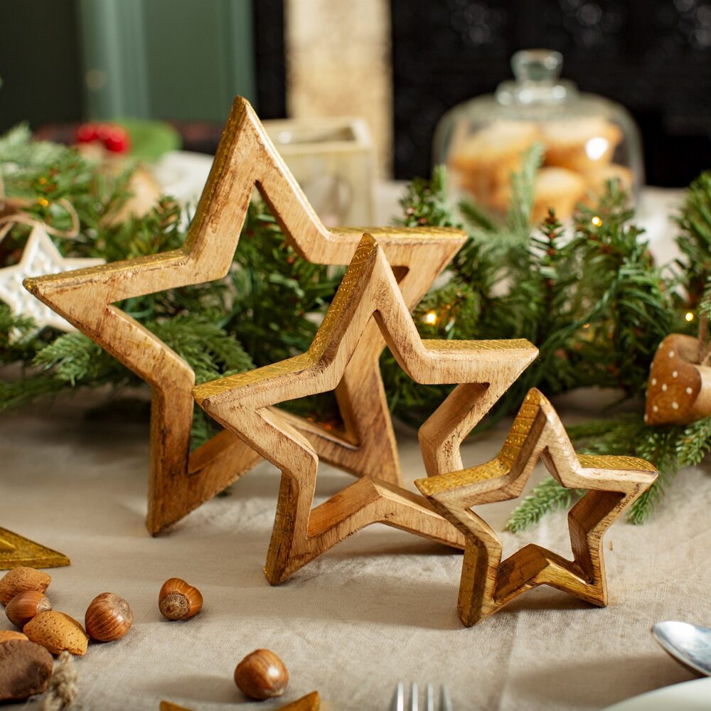 Three wooden star decorations of varying sizes are displayed on a festive table with greenery, nuts, and baubles, creating a cosy holiday atmosphere.