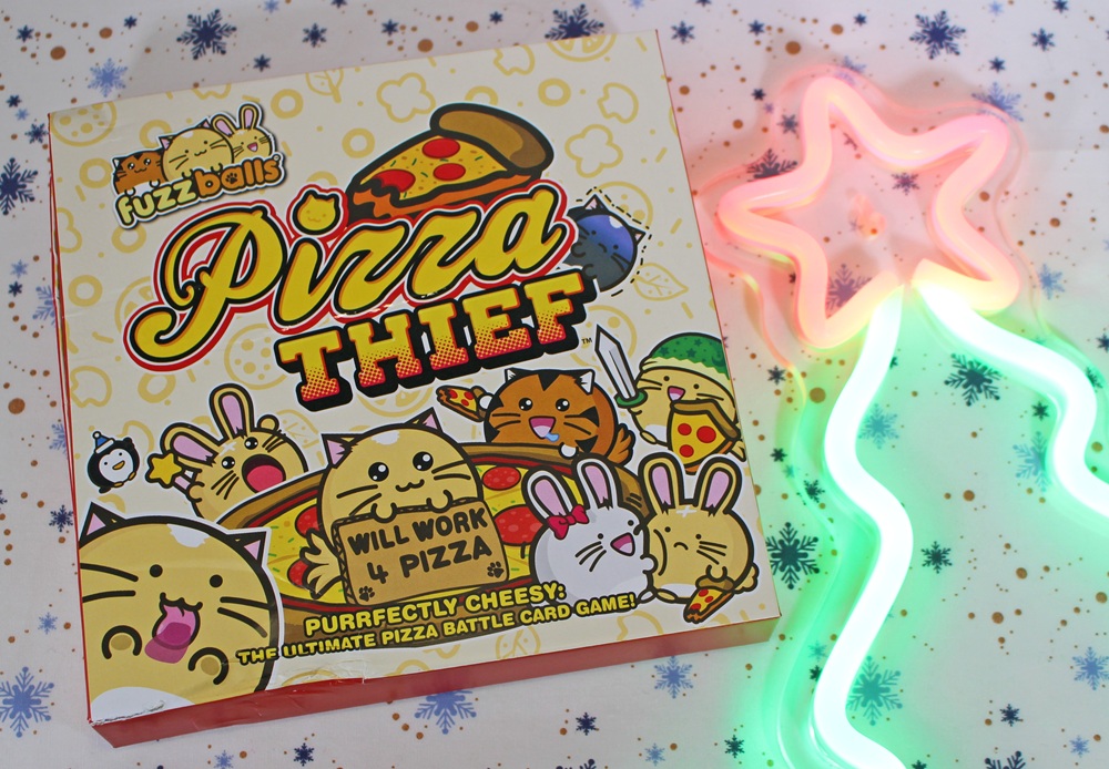 A colourful card game box titled Pizza Thief with cartoon cats and bunnies, pizza slices, and bold yellow text. A neon pink and green star-shaped light is on the right, on a patterned surface.