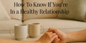 Two people hold hands in front of two coffee mugs on a table, with the text How To Know If You’re In a Healthy Relationship displayed above, against a soft, beige background.
