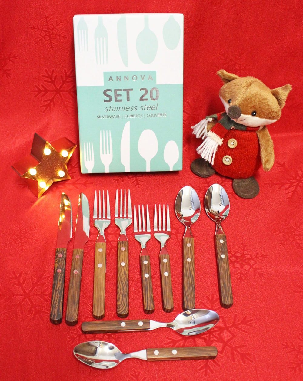 A 20-piece stainless steel cutlery set with wooden handles is displayed on a red cloth, alongside a cute plush fox toy and a small star-shaped light. The set includes knives, forks, tablespoons, and teaspoons.