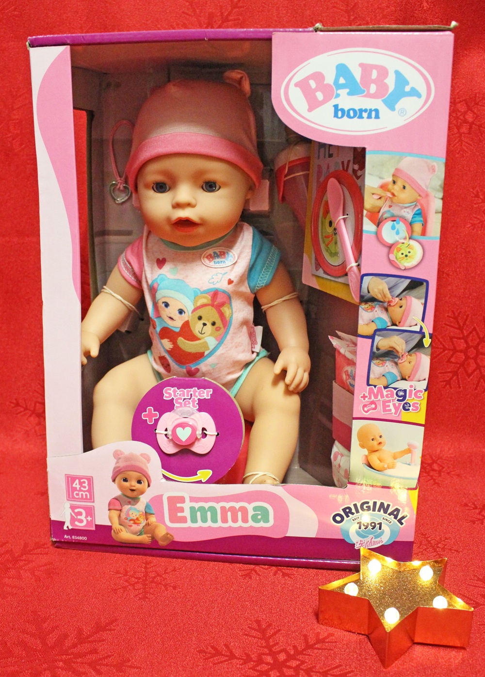 Boxed BABY born doll named Emma, wearing a pink hat and colourful outfit, displayed with accessories. The packaging has playful graphics, Magic Eyes, and Starter set. A lit star-shaped sign reads Original 1991.