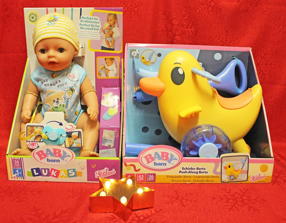 Two boxed BABY born toys displayed on a red background: a baby doll named Lukas wearing a yellow hat and blue bib, and a yellow push-along duck toy with purple wheels. A small lit star decoration sits in front.