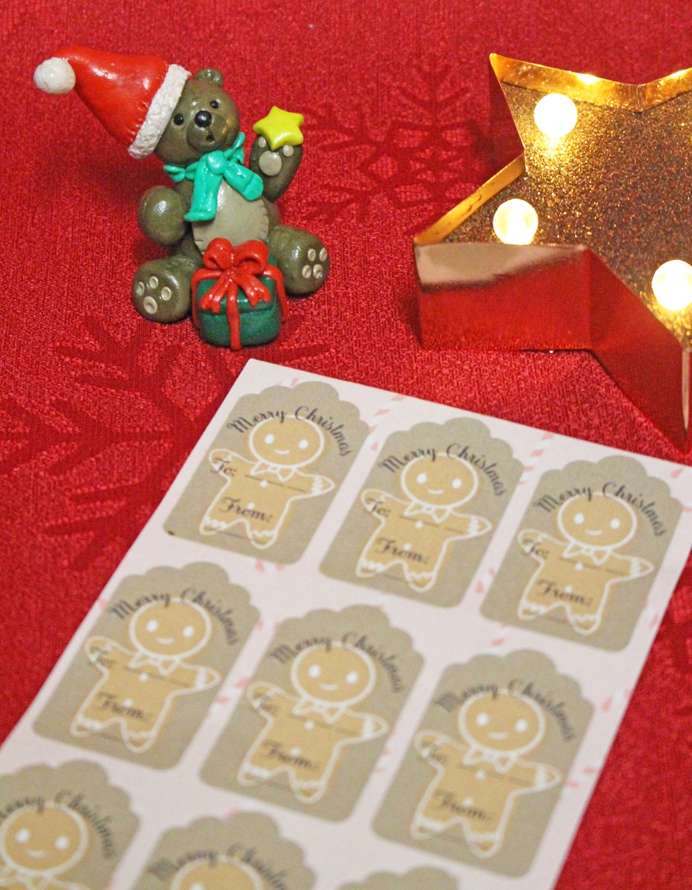A festive scene with a small bear figurine in a Santa hat, a lit star-shaped decoration, and a sheet of Merry Christmas gift tags with gingerbread men on a red textured surface.