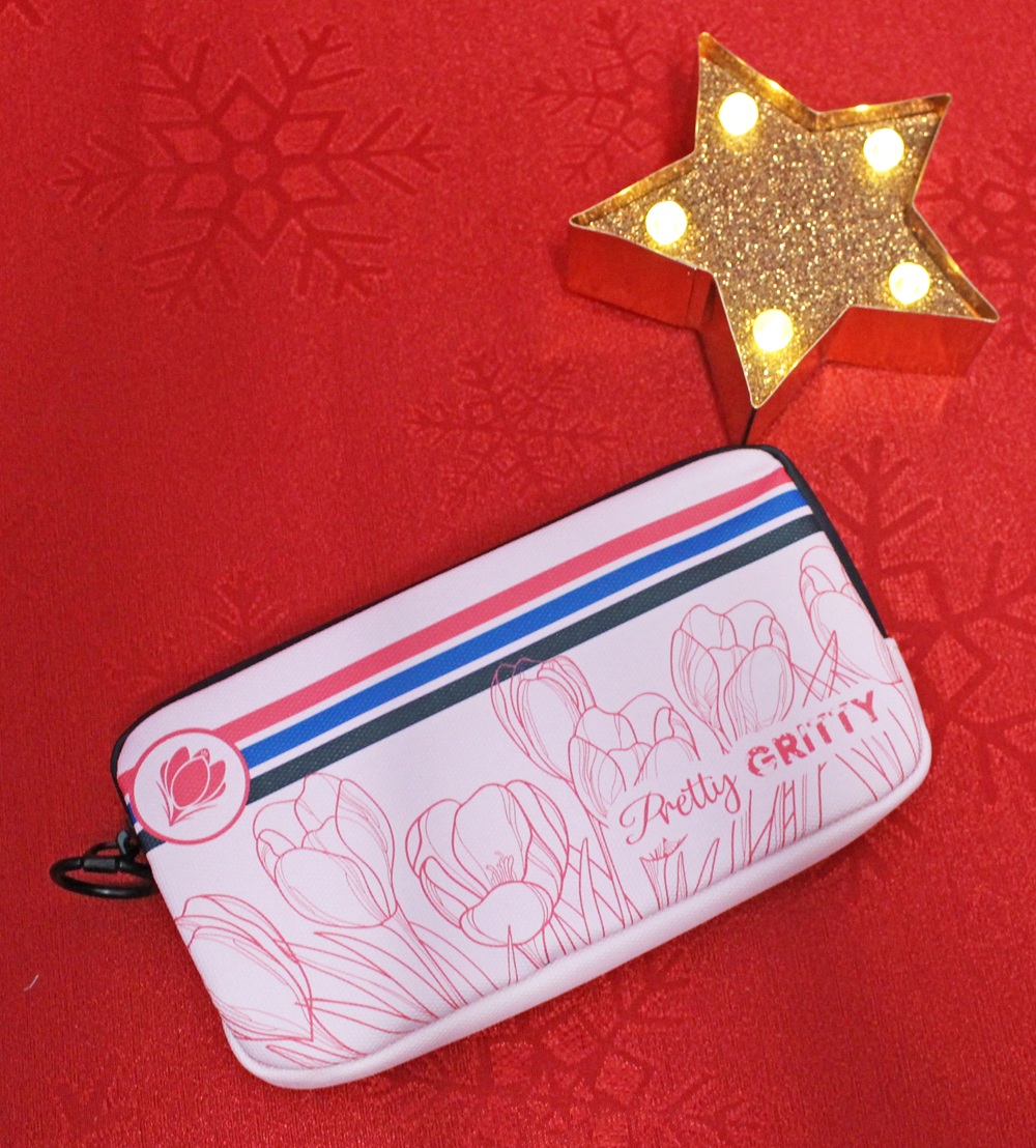 A pink zippered pouch with tulip designs and “Pretty GRITTY” written on it sits on a red surface with snowflake patterns, next to a lit gold star decoration.