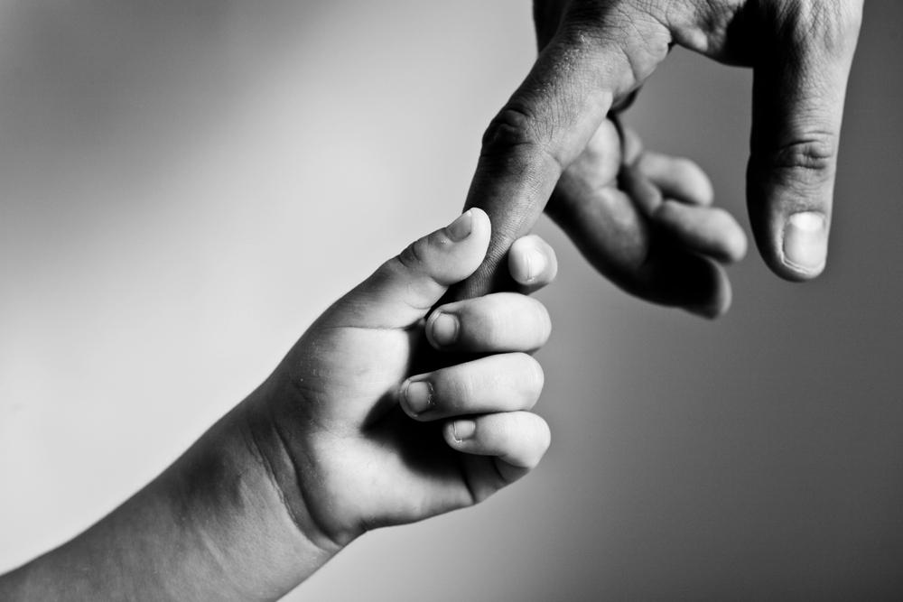 A child’s hand gently holding an adult’s finger against a blurred, light background, symbolising trust, connection, and care.