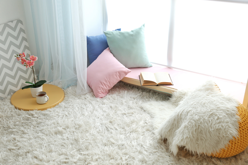 A cosy reading nook with pastel cushions by a window, an open book, a cup of coffee on a small round table, a pink orchid, and a fluffy white rug and blanket creating a soft, inviting atmosphere.