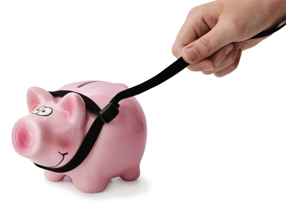 A hand tightens a black belt around a pink piggy bank, symbolising saving money or financial restraint.
