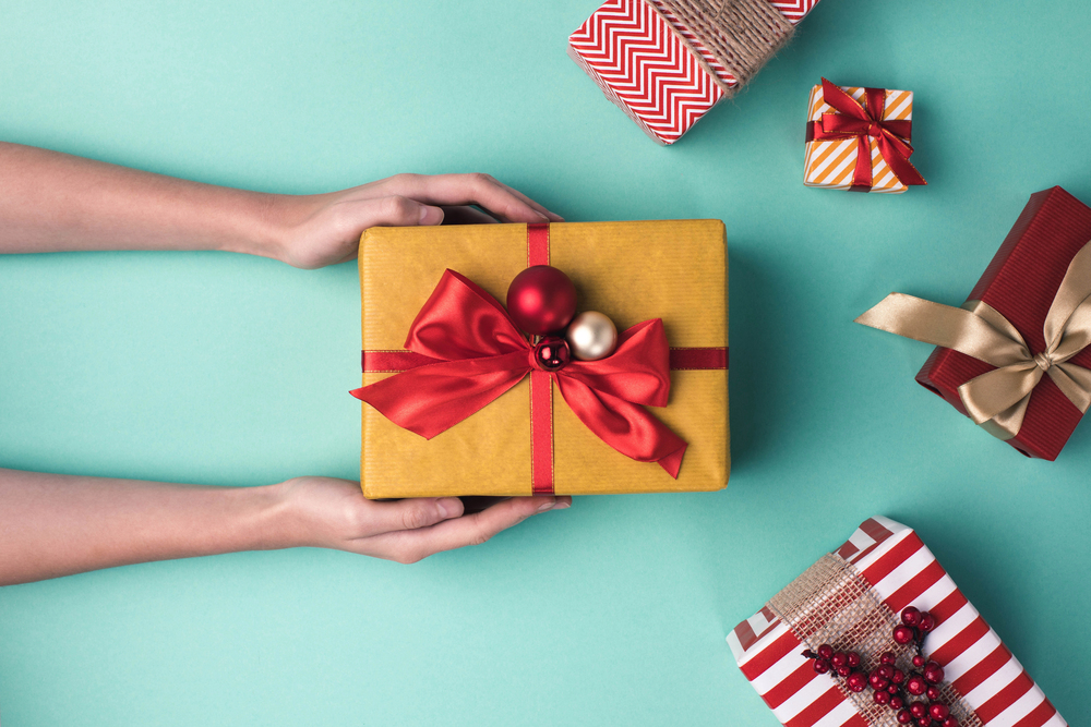 A pair of hands holds a yellow gift box with a red ribbon and ornaments on a turquoise background, surrounded by four other wrapped presents in various colours and patterns.