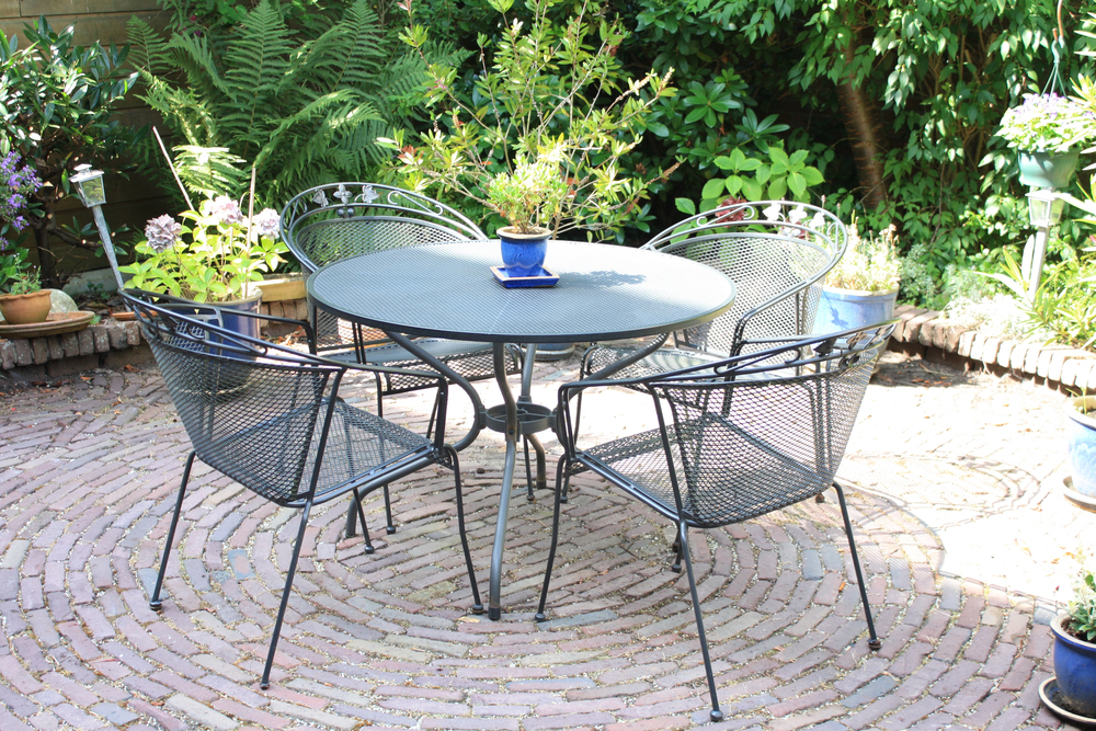 A round metal table with four matching chairs sits on a circular brick patio, surrounded by green plants and potted flowers, with sunlight casting shadows across the scene.