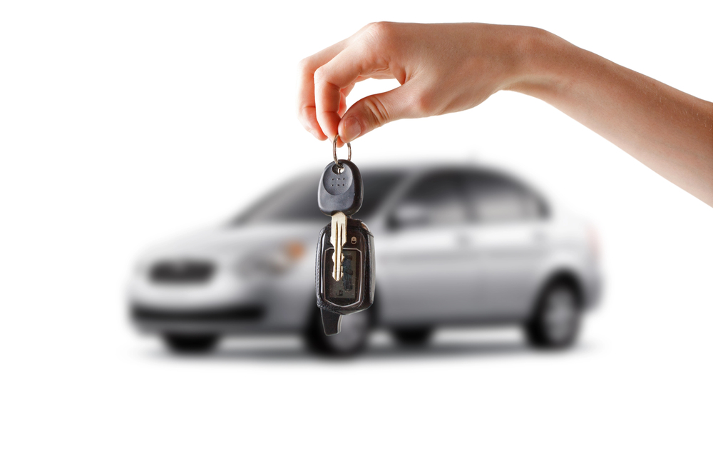 A hand holds up a set of car keys in the foreground, with a blurred silver saloon car in the background on a white surface.
