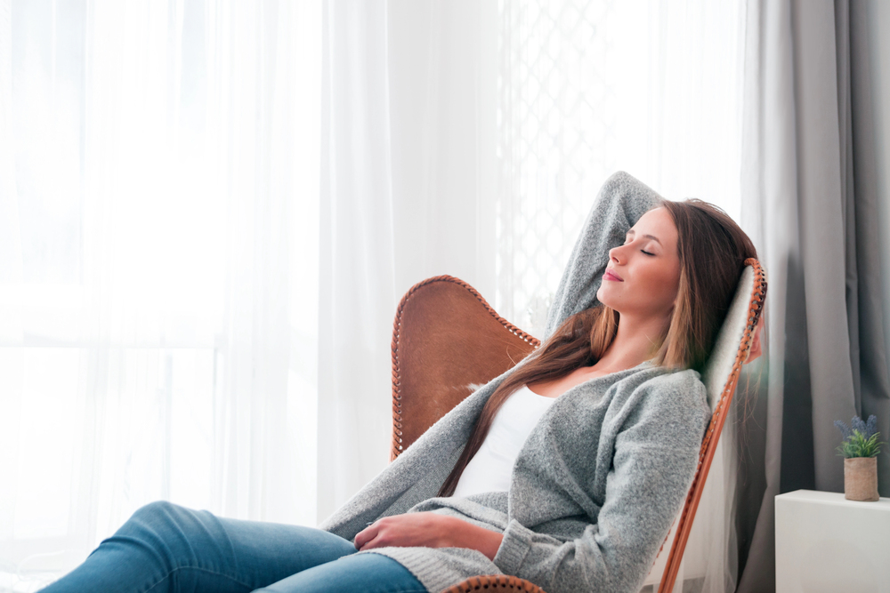 Woman relaxing in a chair with eyes closed, wearing a grey jumper and jeans, sitting by a window with white curtains and soft natural light, appearing peaceful and comfortable.
