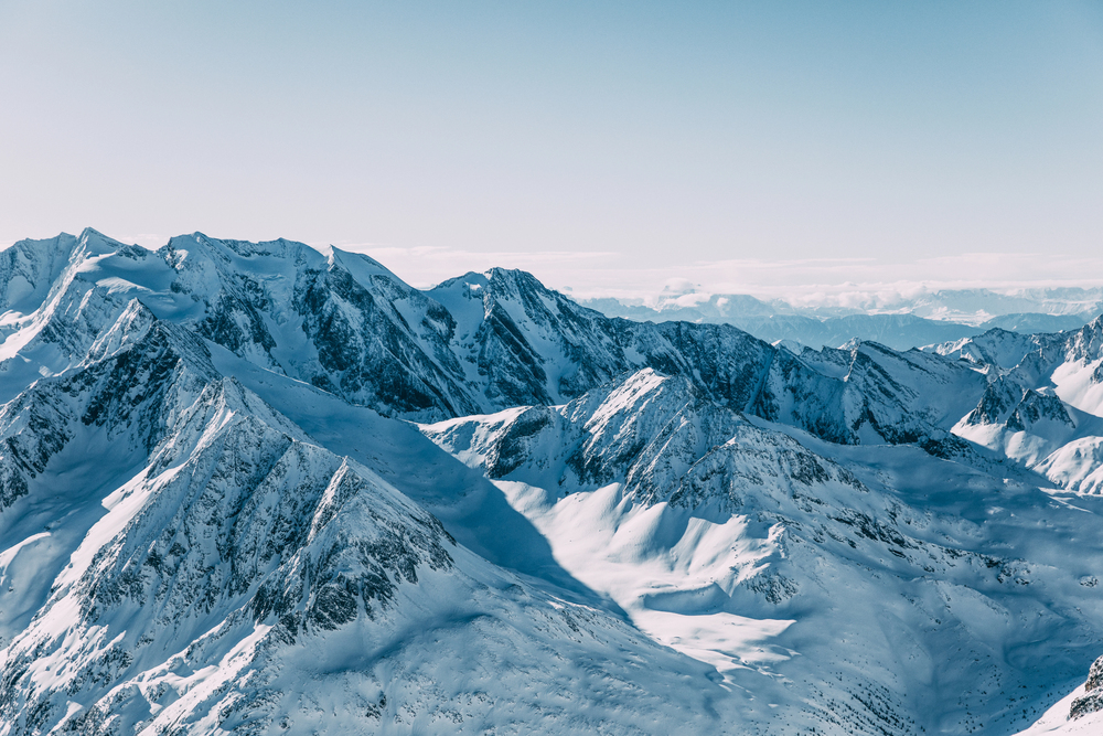 Snow-covered mountain peaks under a clear blue sky, with shadows accentuating the rugged terrain and distant ranges fading into the horizon.