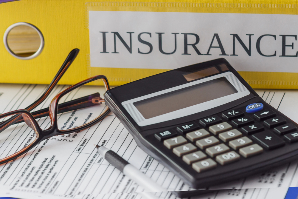 A calculator, spectacles, and a pen rest on insurance forms, with a yellow folder labelled INSURANCE in the background.