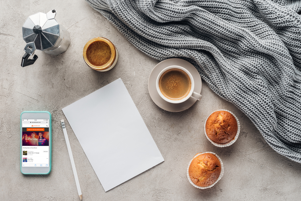A cosy scene with a cup of coffee, two muffins, a blank sheet of paper with a pencil, a moka pot, a jar of coffee, a smartphone with a music app open, and a grey knitted blanket on a grey surface.