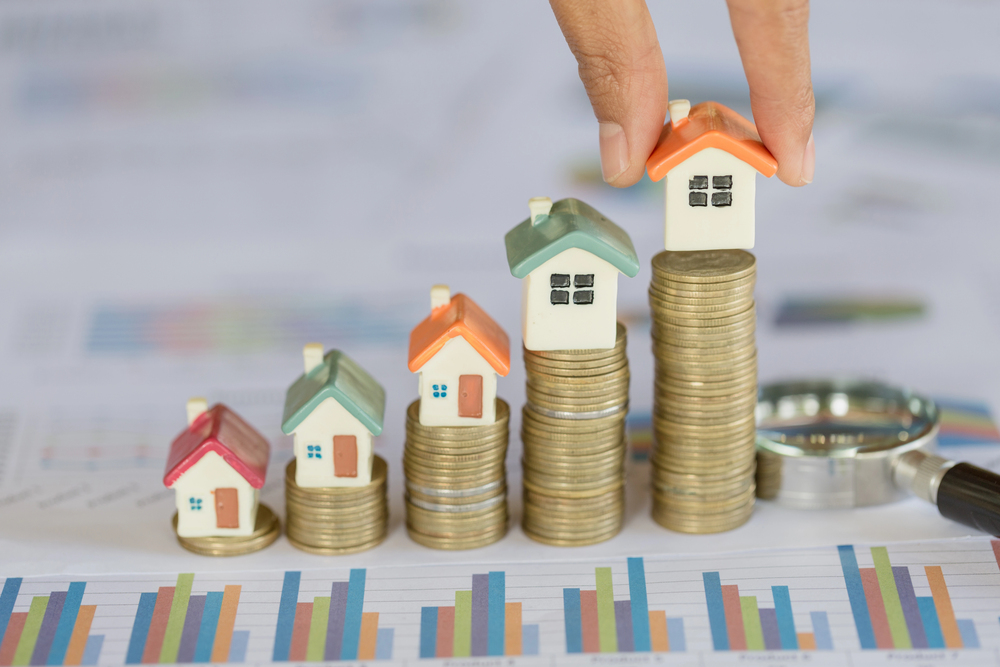 Four stacks of coins, each topped with a small house model, increase in height from left to right. A hand places the tallest house. In the background, blurred charts and a magnifying glass are visible.