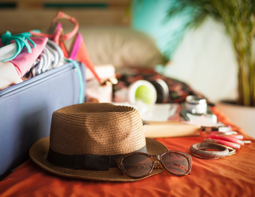 A straw hat and sunglasses rest on an orange bedspread beside an open suitcase with clothes and swimwear, suggesting preparations for a holiday. A blurred plant and other travel items are visible in the background.