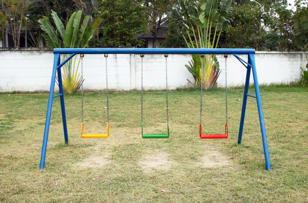 A blue metal swing set with three empty swings—yellow, green, and red—on a grassy garden, with trees and a white wall in the background.