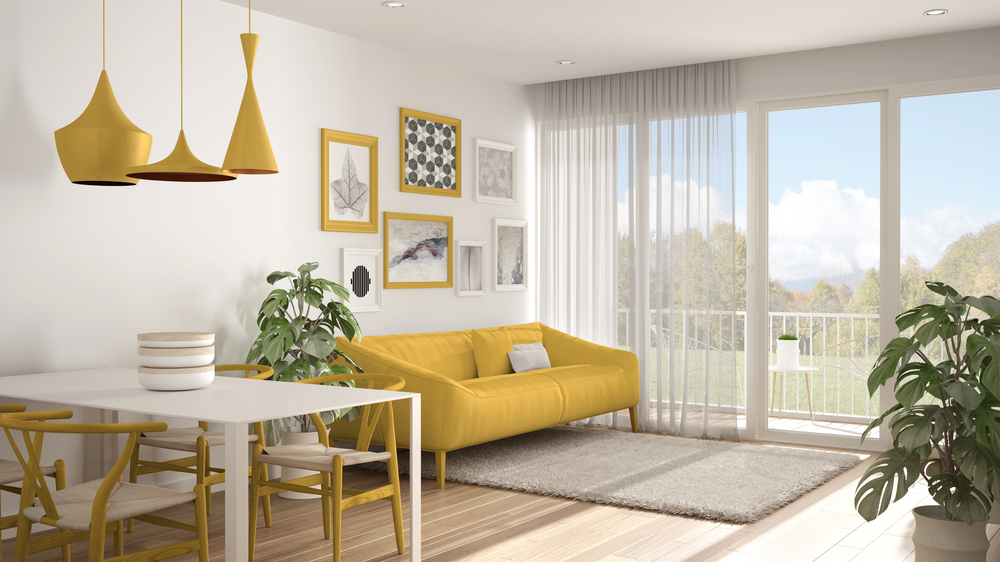 Bright modern living room with large windows, a yellow sofa, yellow hanging pendant lights, wall art, green plants, a white dining table with chairs, and a grey rug on wooden floors.